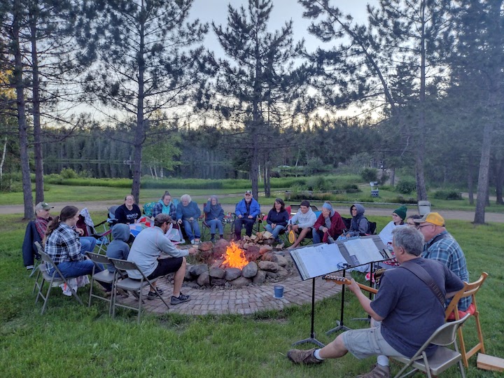 people gathered around a fire circle, a couple guys playing guitar, lake in the background