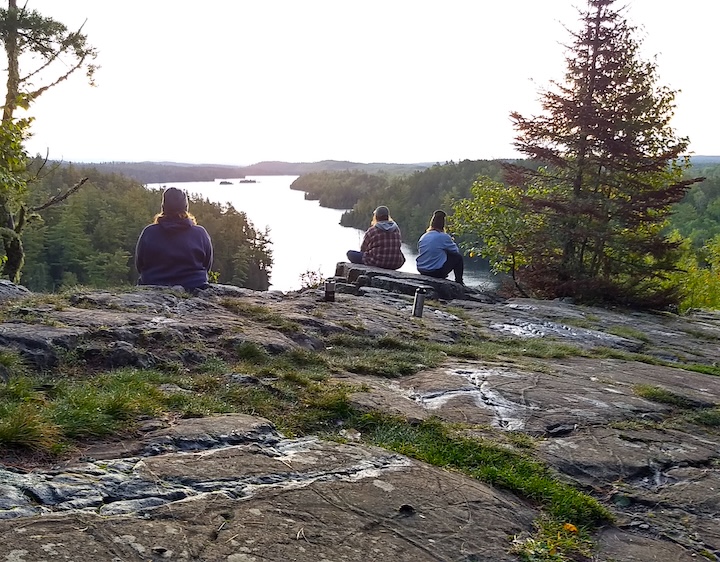 three hikers sit at an overlook of a lake at sunrise