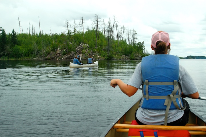 a woman sits in the bow of a canoe, with another canoe in the background with two women