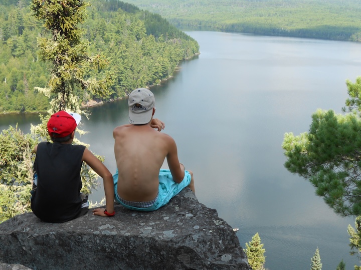 two boys sit on a rock overlooking Rose Lake in the Boundary Waters