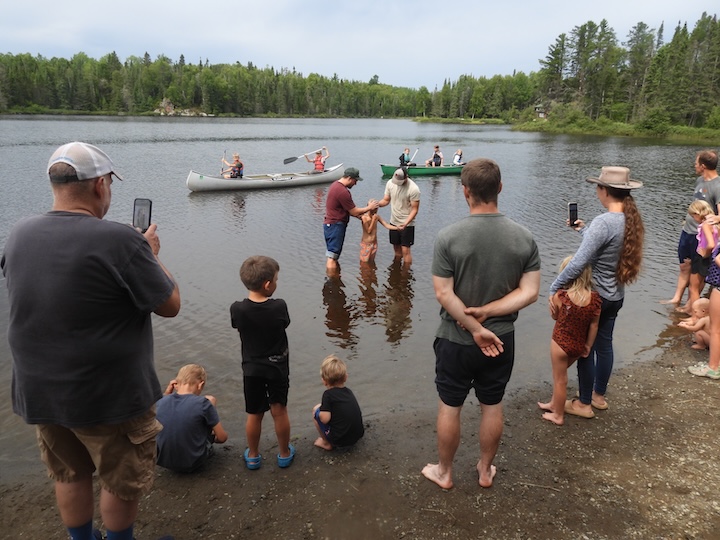 two dads ready to baptize a young boy in Bow Lake at Okontoe while several watch from the shore and in canoes