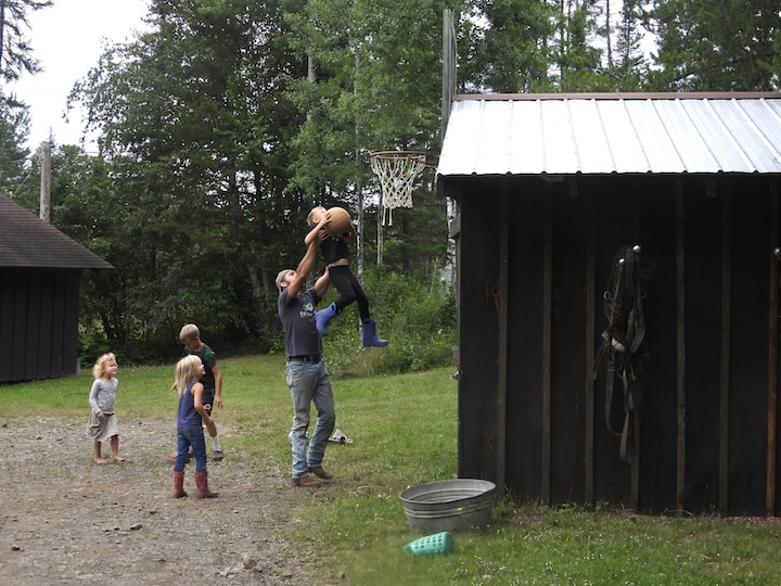 an adult man holds up a young boy so he can shoot the basketball through the hoop, with a few others young kids surrounding them