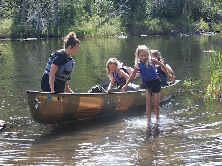 a couple of moms and two young girls get out of a canoe at a portage