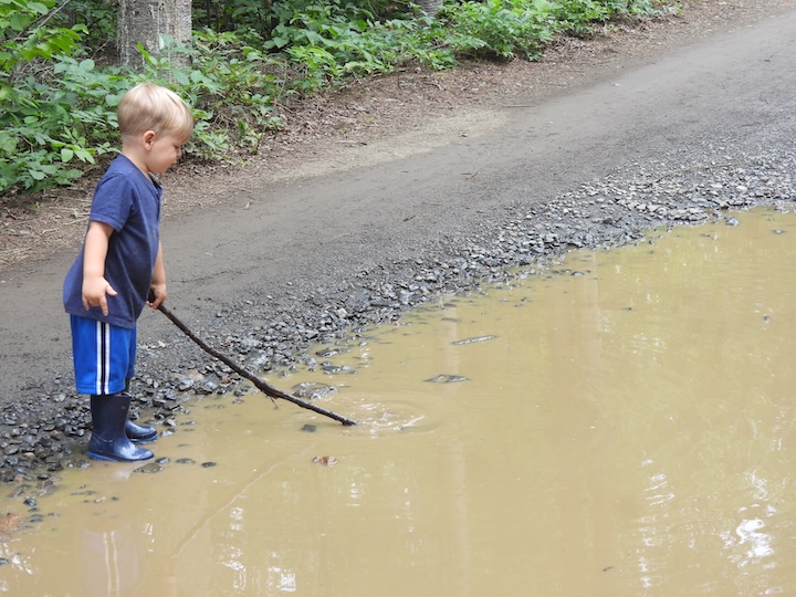 a toddler plays with a stick in a large mud puddle