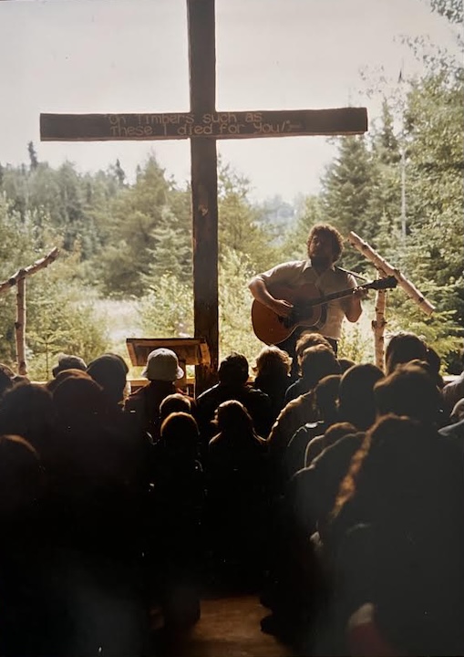 a man leads worship before a cross in a northwoods chapel